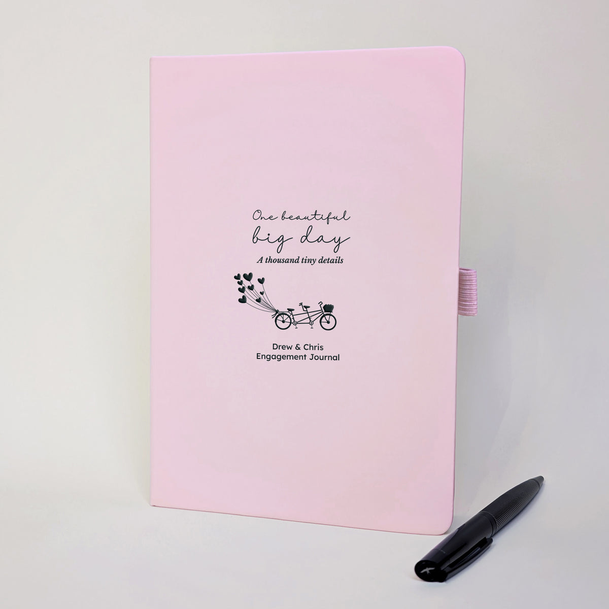 Pink personalised engagement journal with black tandem bicycle and heart balloons, names Drew and Chris, “One beautiful big day, a thousand tiny details” and black metal pen.