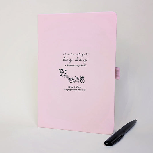Pink personalised engagement journal with black tandem bicycle and heart balloons, names Drew and Chris, “One beautiful big day, a thousand tiny details” and black metal pen.