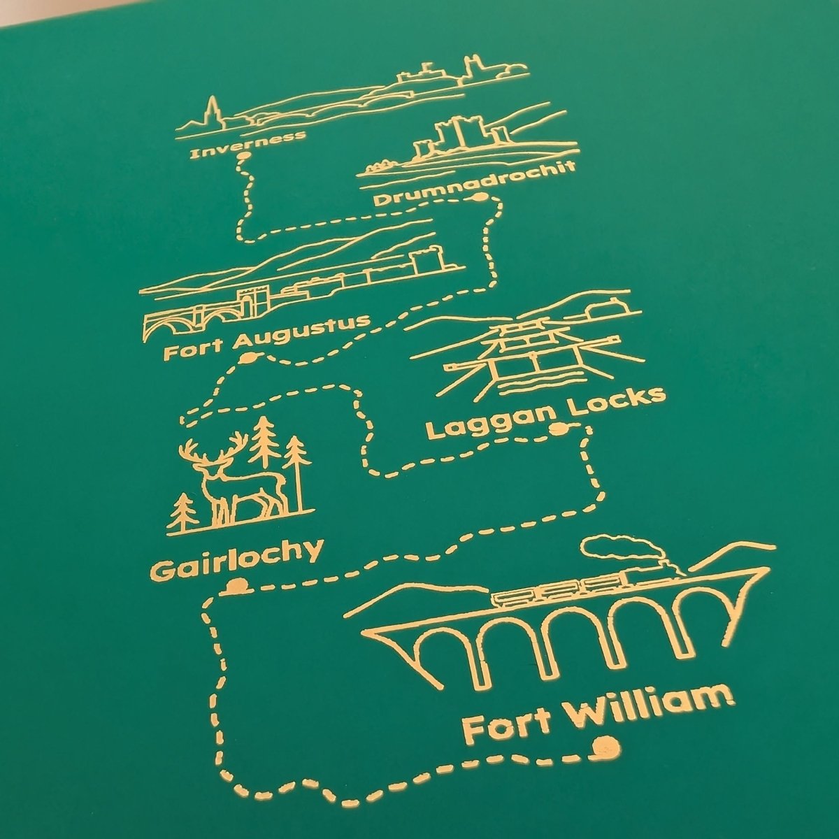 Close-up of detailed gold foiled Scottish Highlands hiking route on a green journal, featuring a stag, landmarks and destination names.