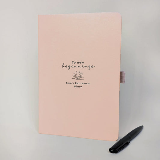 Personalised blush retirement diary with “To new beginnings”, sunrise illustration and custom name, paired with a black metal pen.