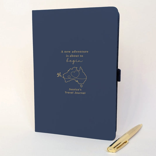 Personalised navy travel journal with gold foil Australia map, aeroplane flight path and custom name, shown with a matching gold metal pen.
