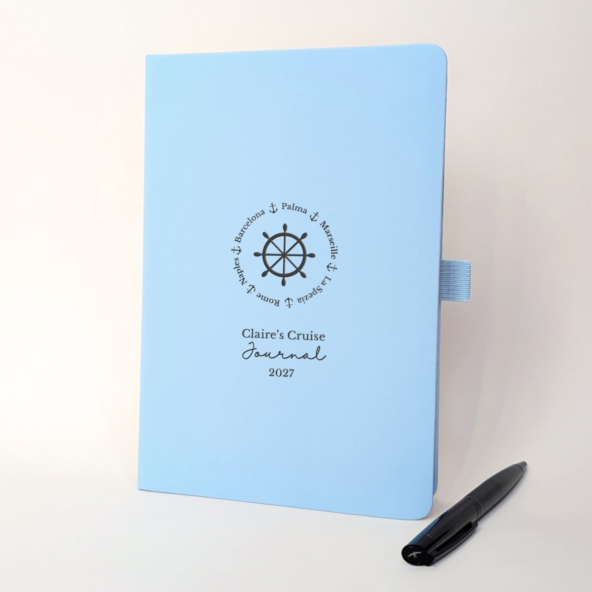 Personalised light blue cruise journal with custom cruise ports arranged around a ship’s wheel, name, year and black metal pen.