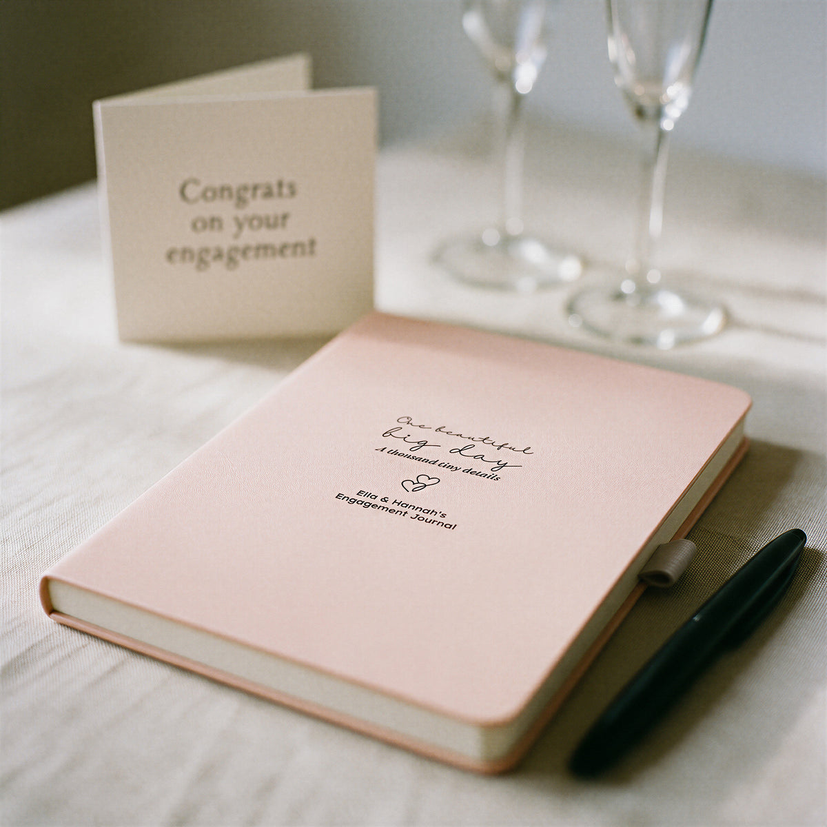 Blush personalised engagement journal with black pen styled on a table with champagne flutes and an engagement card.