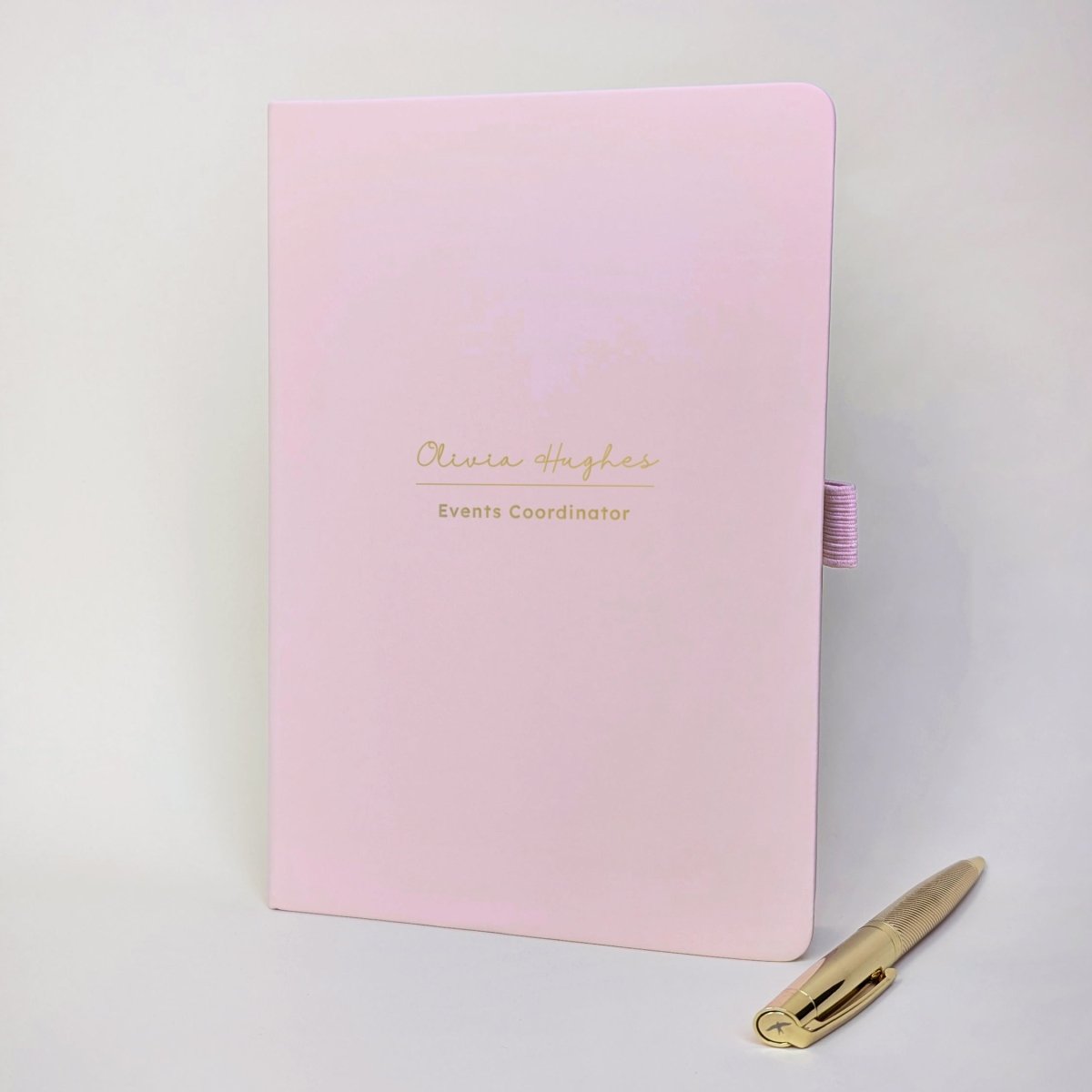 Pink personalised A5 work journal with Olivia Hughes and Events Coordinator on the cover in gold, with matching gold pen. Personalised notebook gift for a colleague, employee or new job.