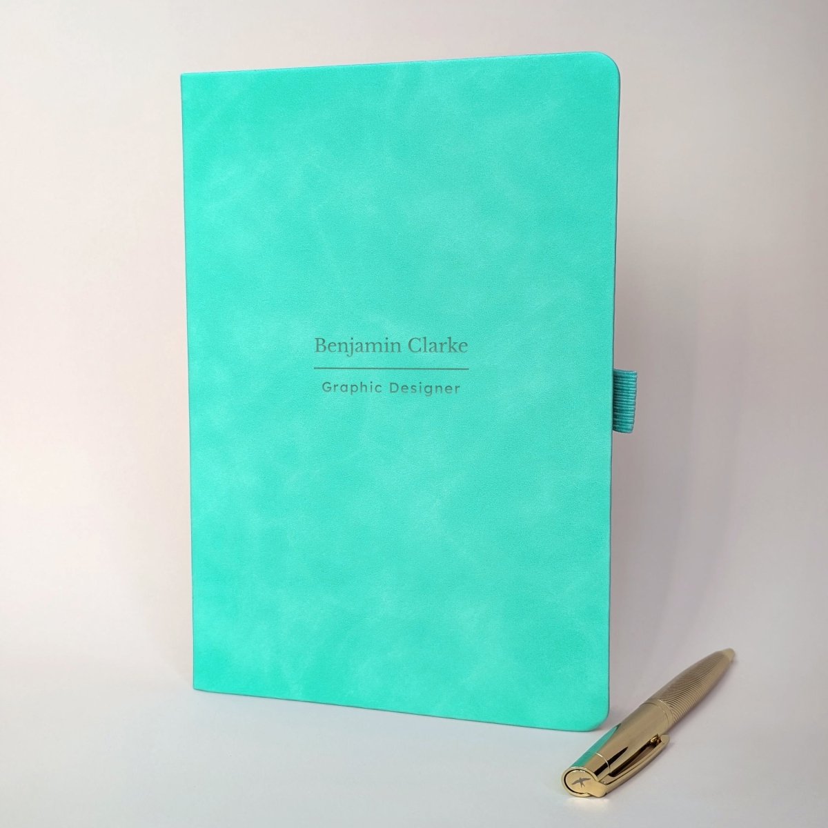 Turquoise personalised A5 journal with Benjamin Clarke and Graphic Designer on the cover, shown with a metal pen. Personalised name and job title notebook for work, a new role or colleague gift.