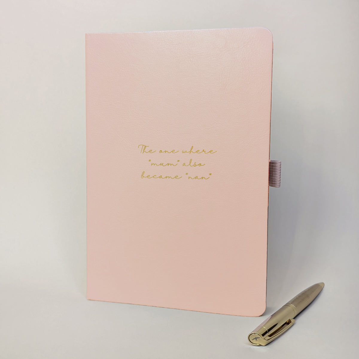 Blush pink personalised journal and gold pen with the cover wording “The one where mum also became nan”, shown as a keepsake gift idea for a new grandmother or family milestone.