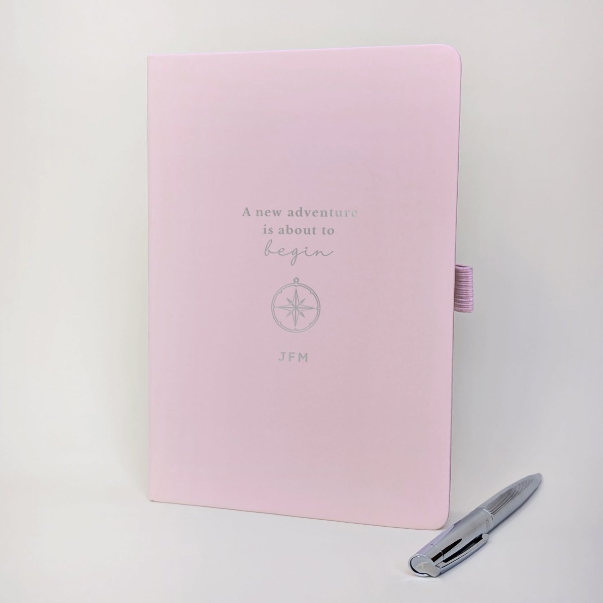 Personalised pink A5 travel journal with compass illustration and custom initials, shown with a silver metal ballpoint pen.