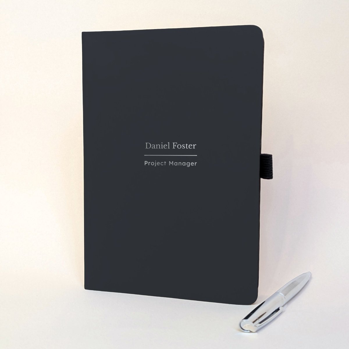 Personalised black A5 work journal with Daniel Foster and Project Manager on the cover, shown with a silver metal pen. A thoughtful personalised gift for a colleague, new job, promotion or work milestone.