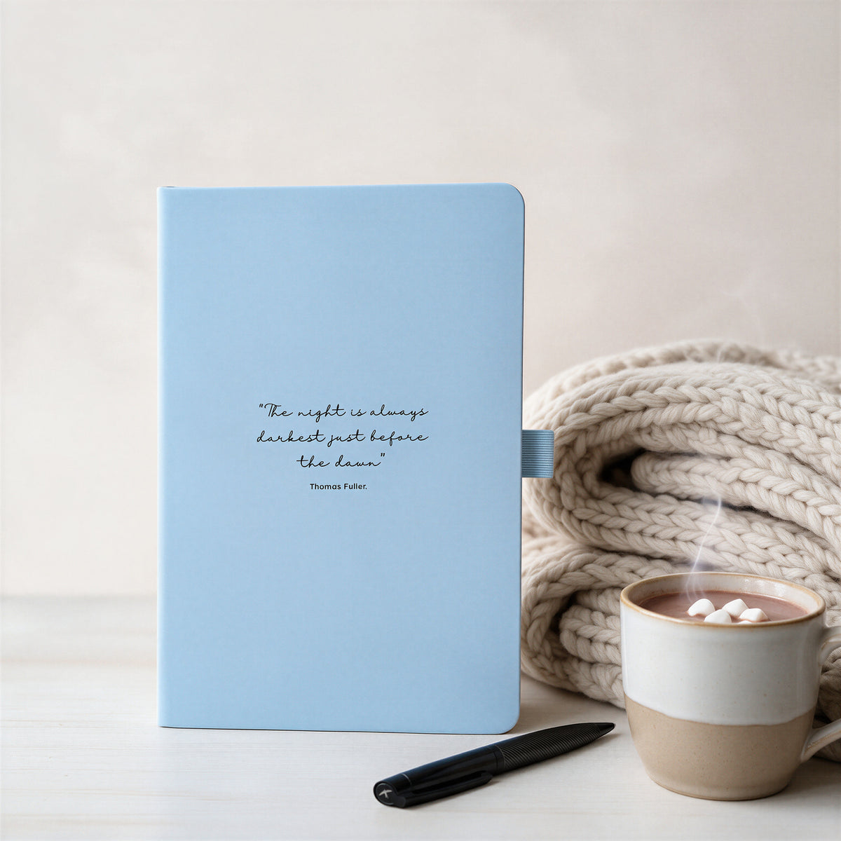 Pale blue personalised quote journal with black pen, chunky knit blanket and hot drink in a cosy reflective setting.