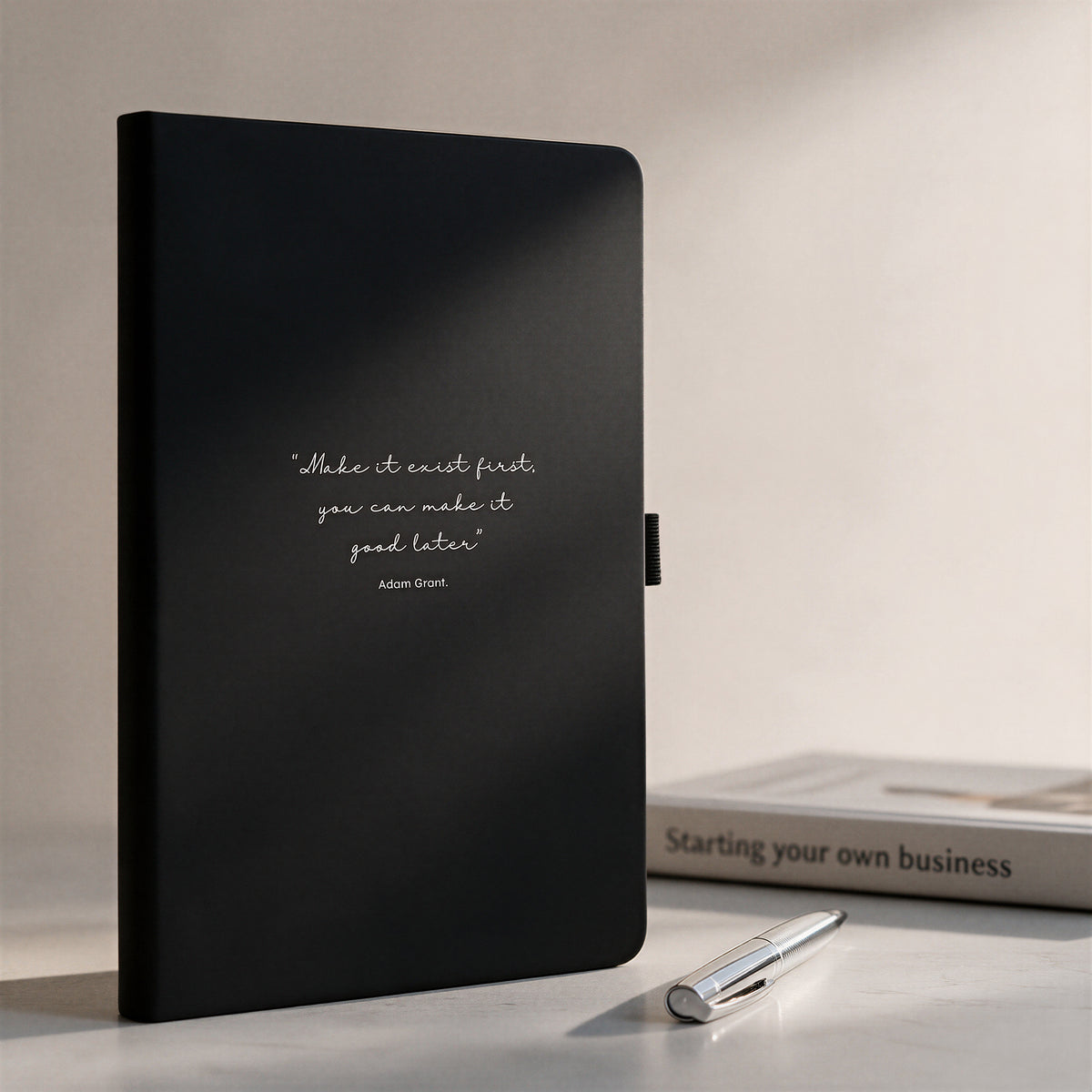 Personalised black quote journal with silver pen styled beside a new business book, showing a custom inspirational cover quote.