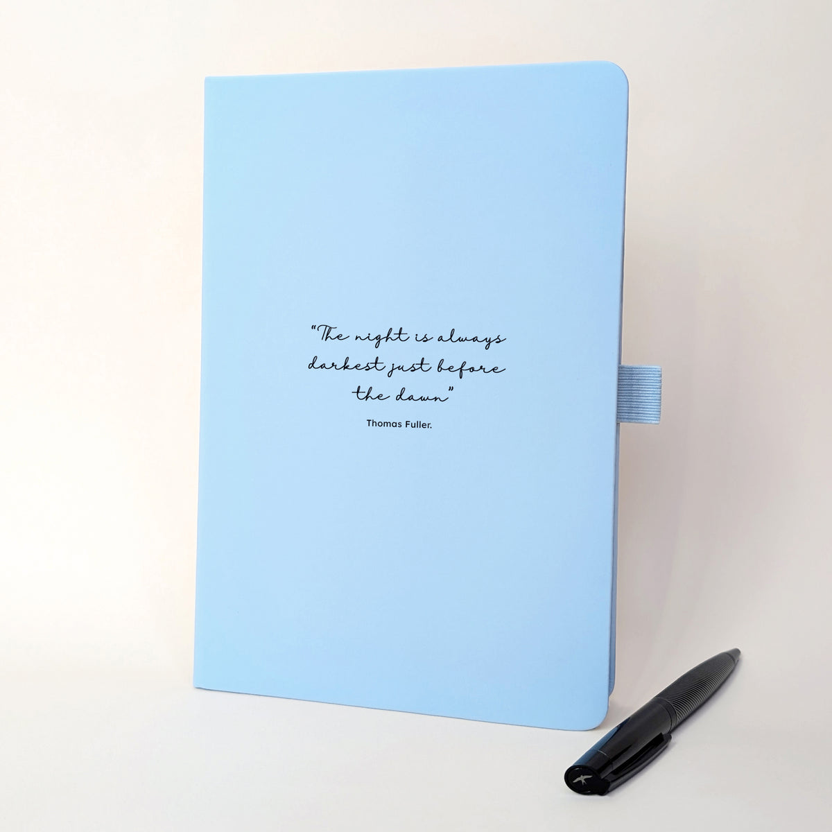 Light blue personalised quote notebook with black text reading “The night is always darkest just before the dawn” by Thomas Fuller, shown with a black pen.