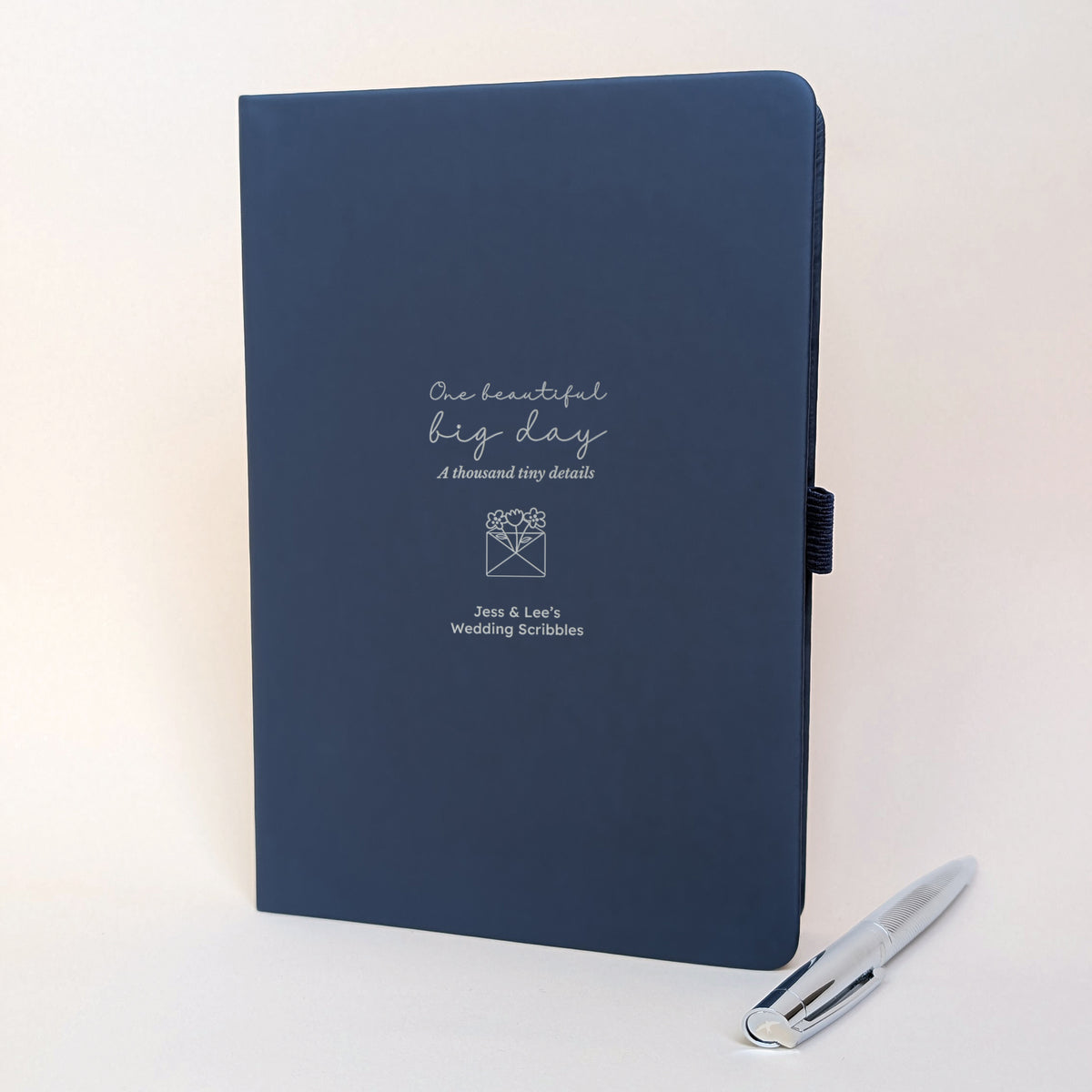 Navy personalised wedding planning journal with silver flower invitation design, “One beautiful big day, a thousand tiny details”, names Jess and Lee, and matching silver metal pen.