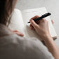 Person writing wedding venue options in a personalised lined journal using a black pen.
