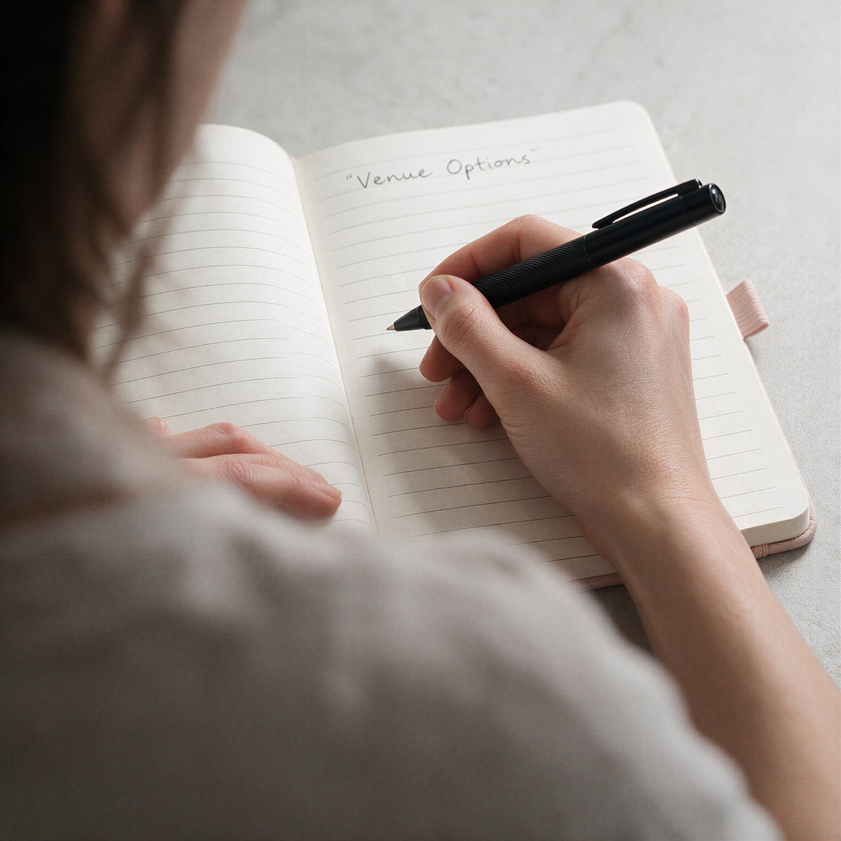 Person writing wedding venue options in a personalised lined journal using a black pen.